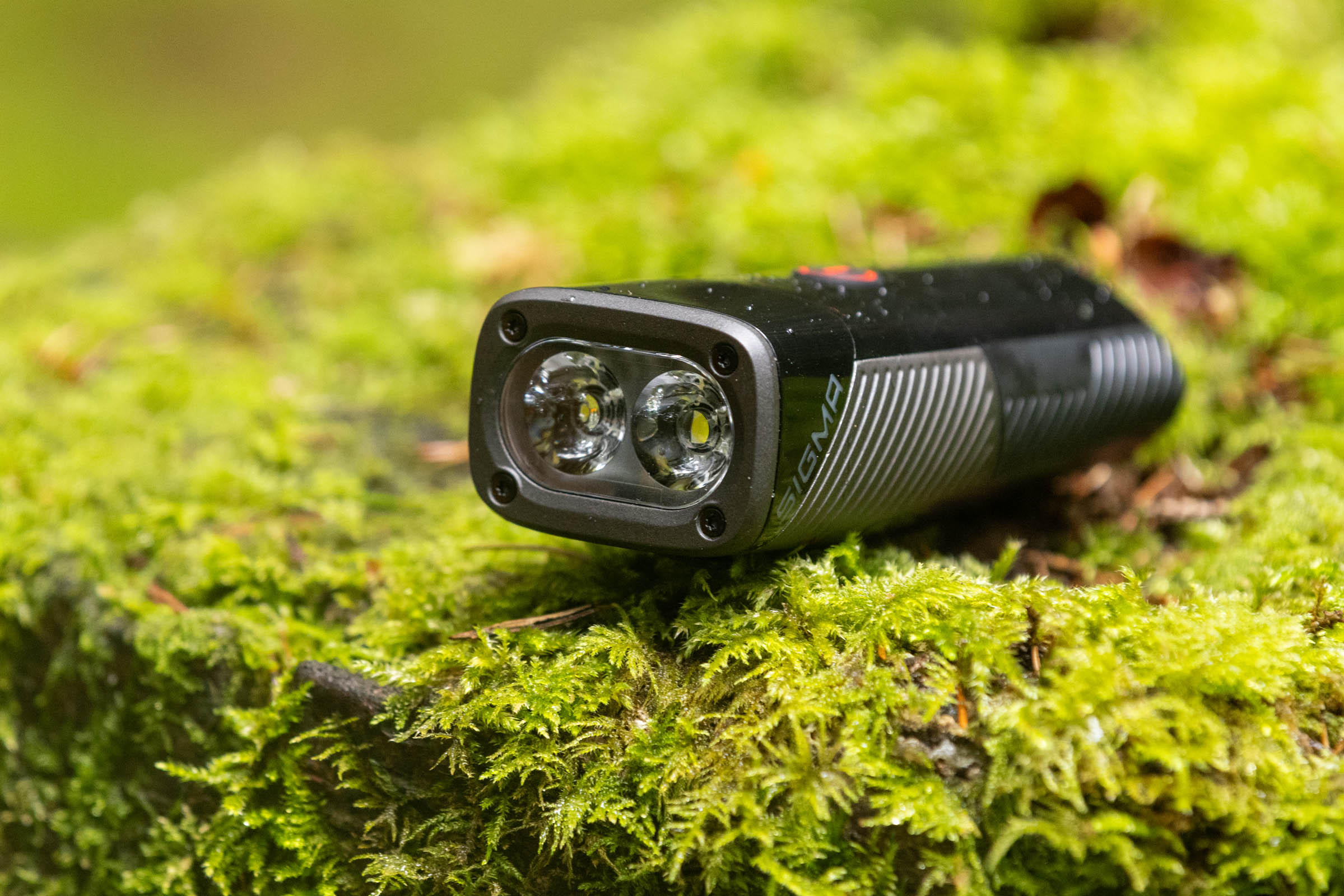 Close-up of a Sigma Buster 1600 mountain bike front light showing its two LED lenses while resting on moss.