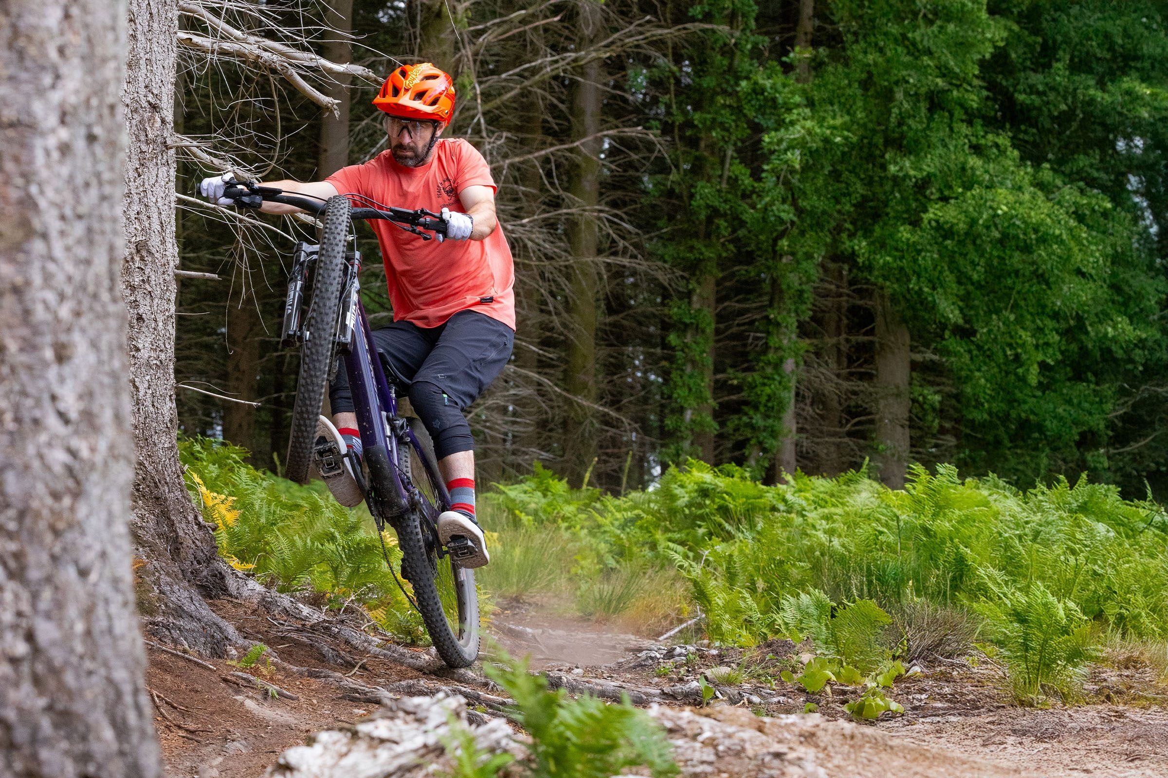 Rider in red jersey, red helmet and black shorts manualling over a root spread on a Santa Cruz Tallboy.