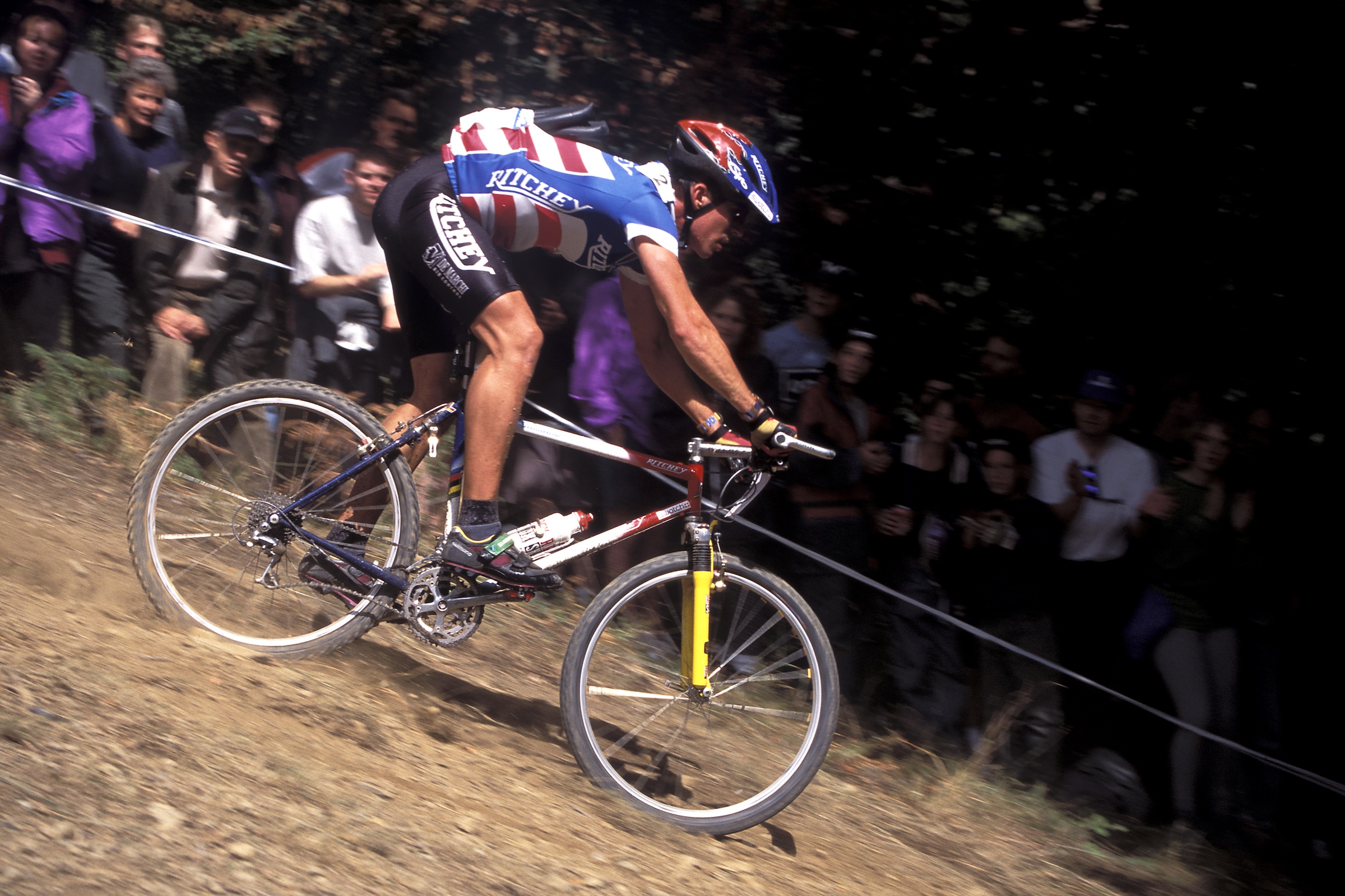 Mountain biker racing a Ritchey mountain bike on a dusty downhill section in front of spectators.