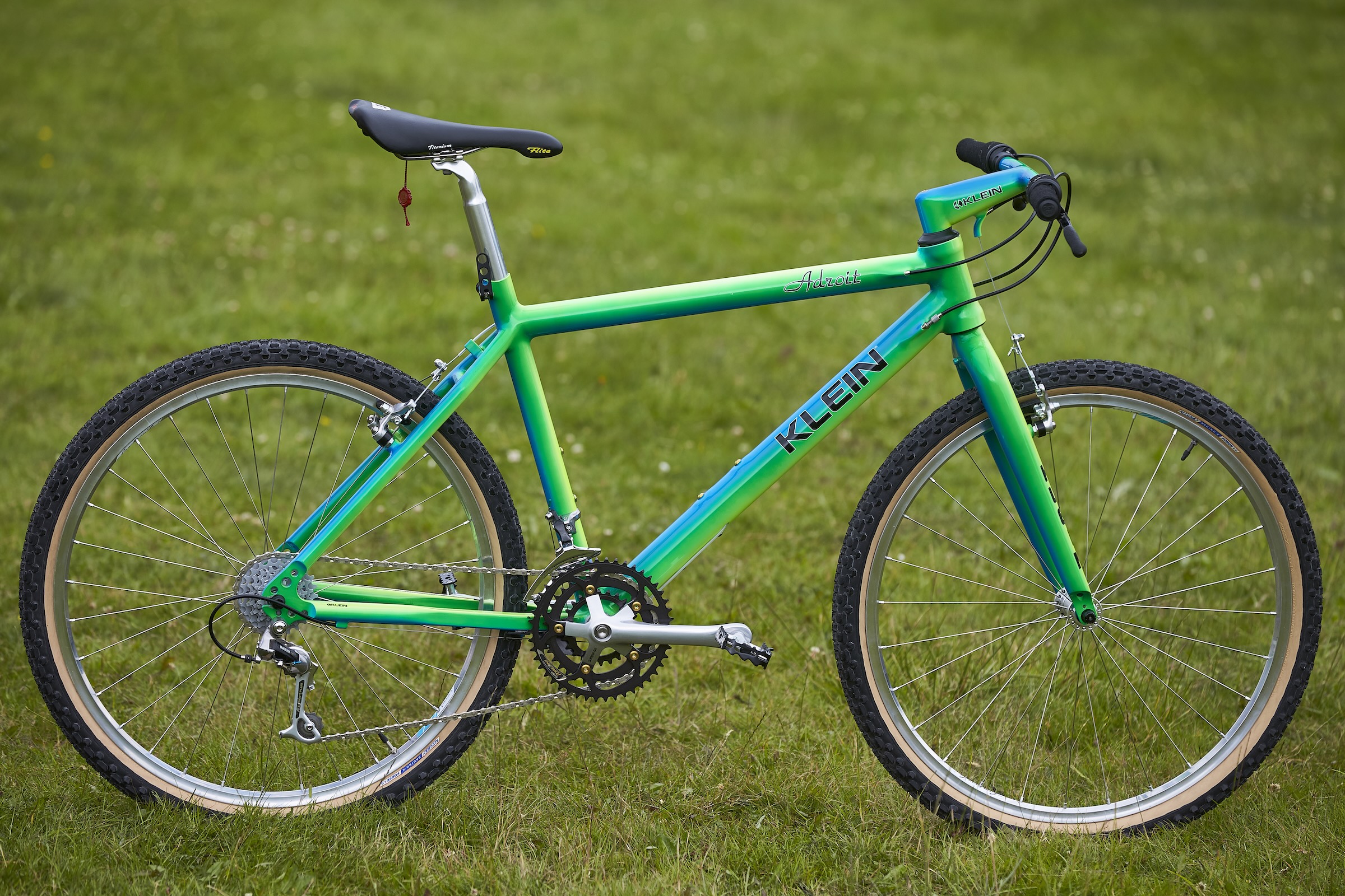 Green and blue Klein Adroit hardtail mountain bike photographed side-on in a grassy field.