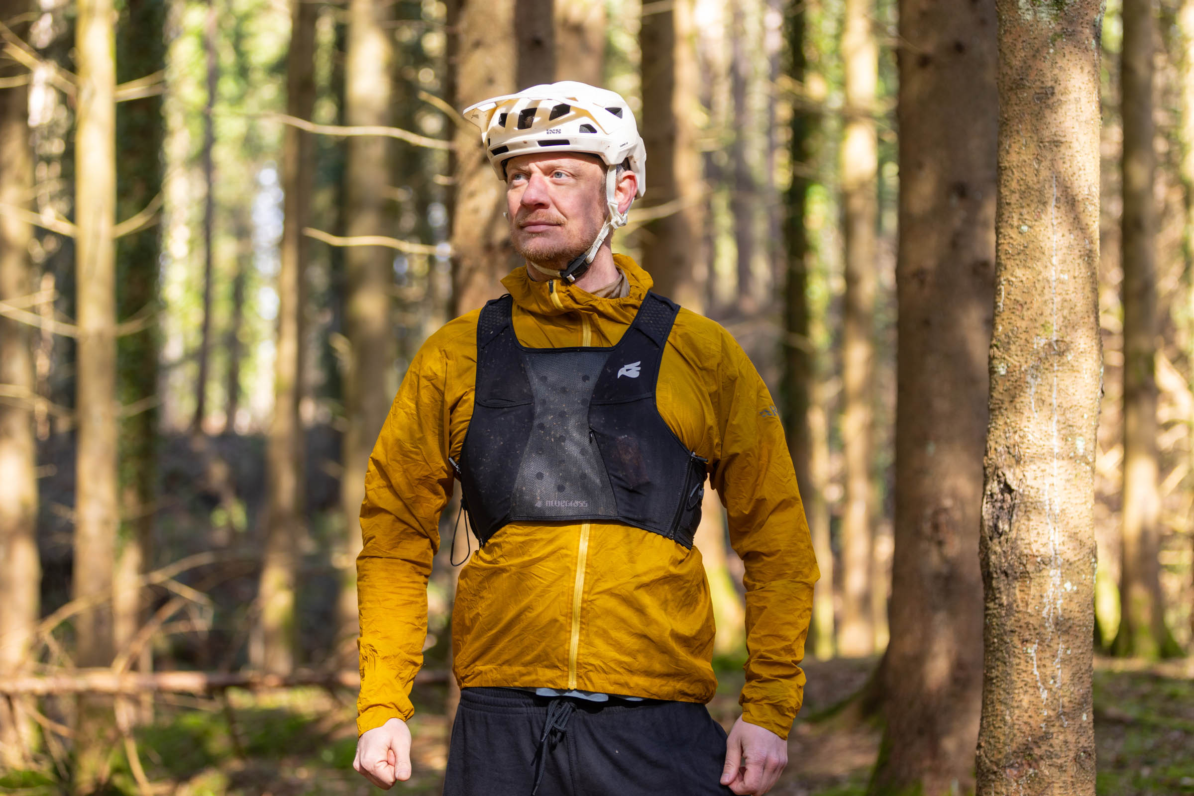 Mountain biker wearing a black Bluegrass Venture Vest over a yellow jacket, standing in a sunlit forest.