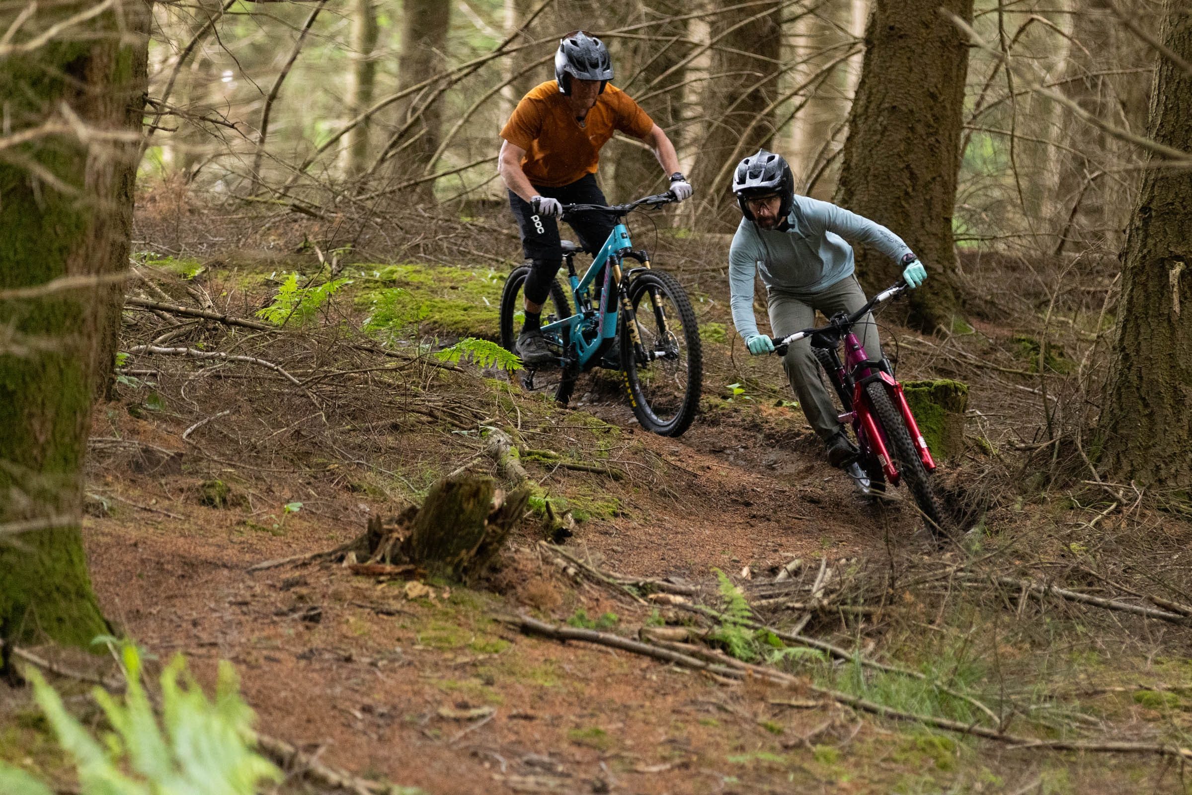 Two riders following one after the other around a right hand turn in dense woodland.