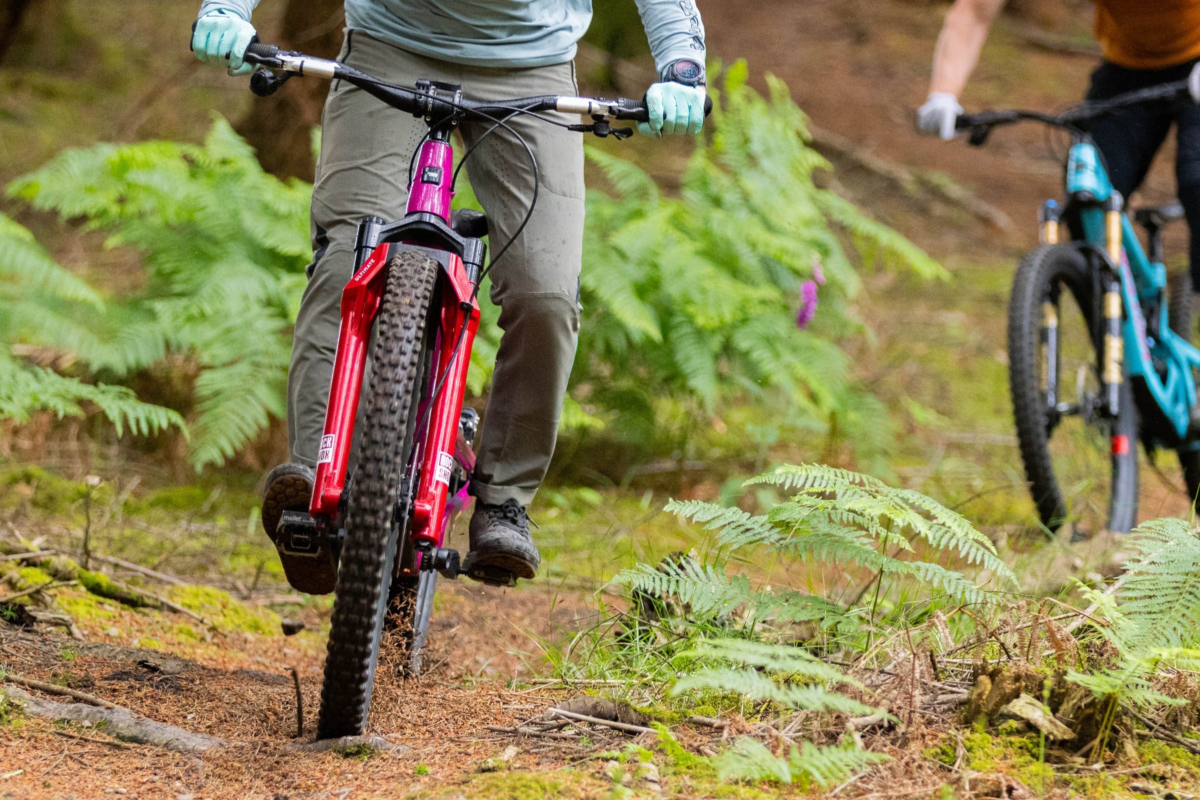 Rider compressing suspension fork while on singletrack trail, followed by another ride close behind.