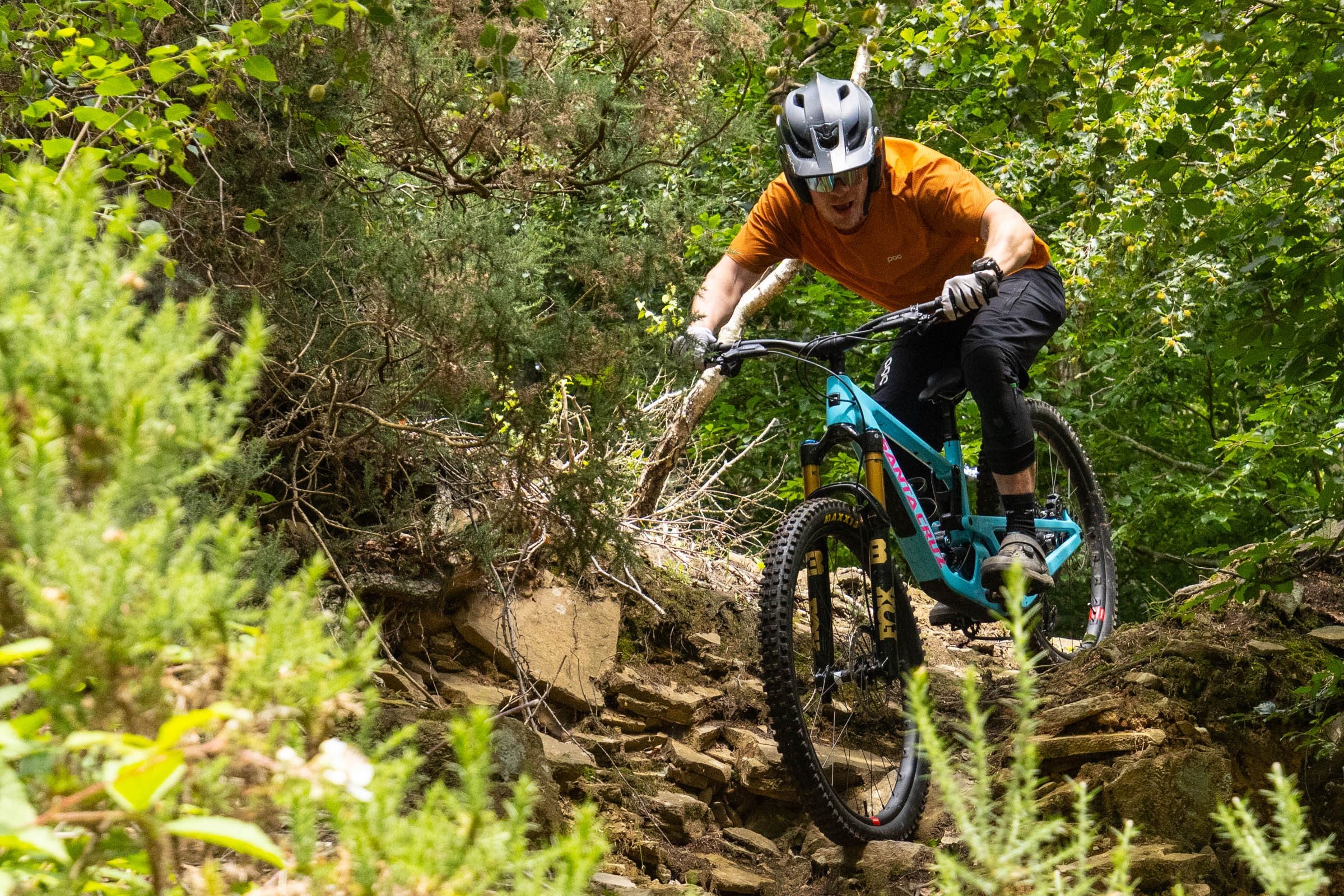 Rider tackling a slabby, enclosed trail with rocks underneath the tyres and trees flanking him either side.