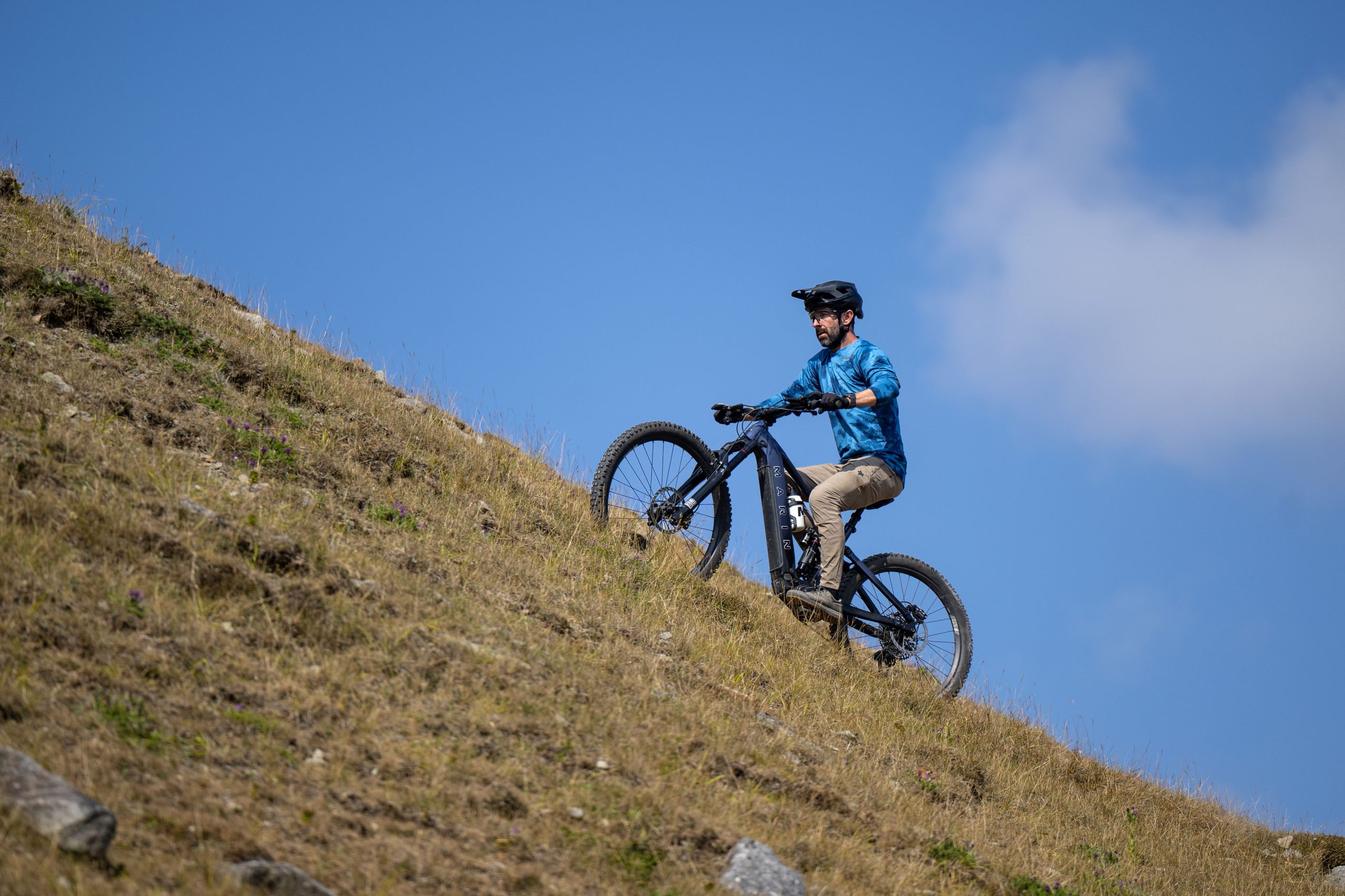 Rider climbs a steep grassy hillside on the dark blue Marin Alpine Trail E XR eMTB with the front wheel lifting