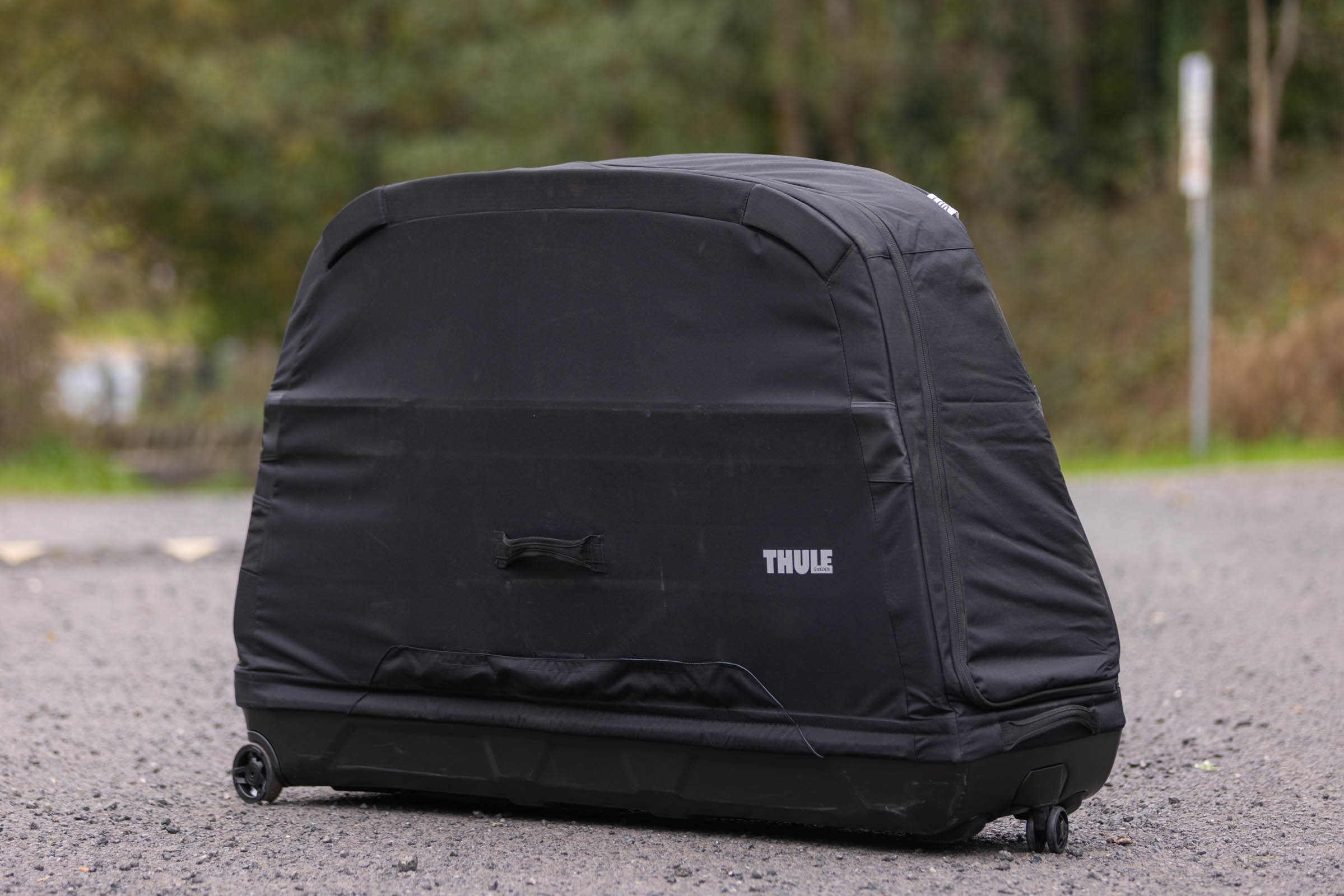 Black Thule RoundTrip MTB bike travel case standing upright on gravel, showing its fabric upper, rigid base, carrying handle and wheels.