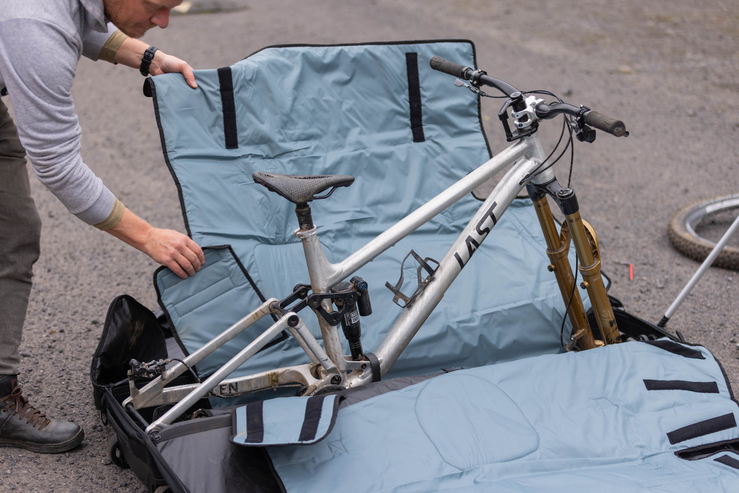 Person positioning a blue padded protective panel around a silver Last mountain bike inside an open Thule RoundTrip MTB travel case.