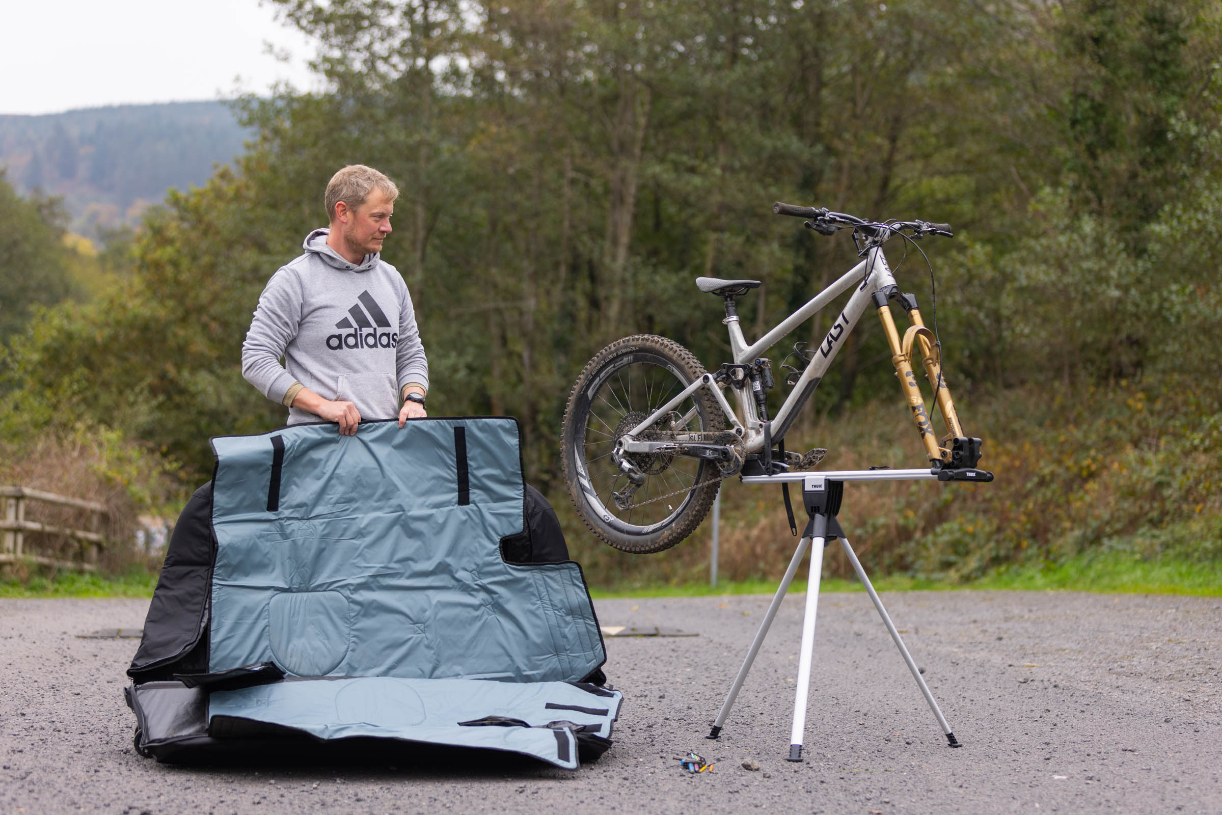 Man standing beside an open Thule RoundTrip MTB travel case, with a silver Last mountain bike mounted on its integrated three-legged workstand.