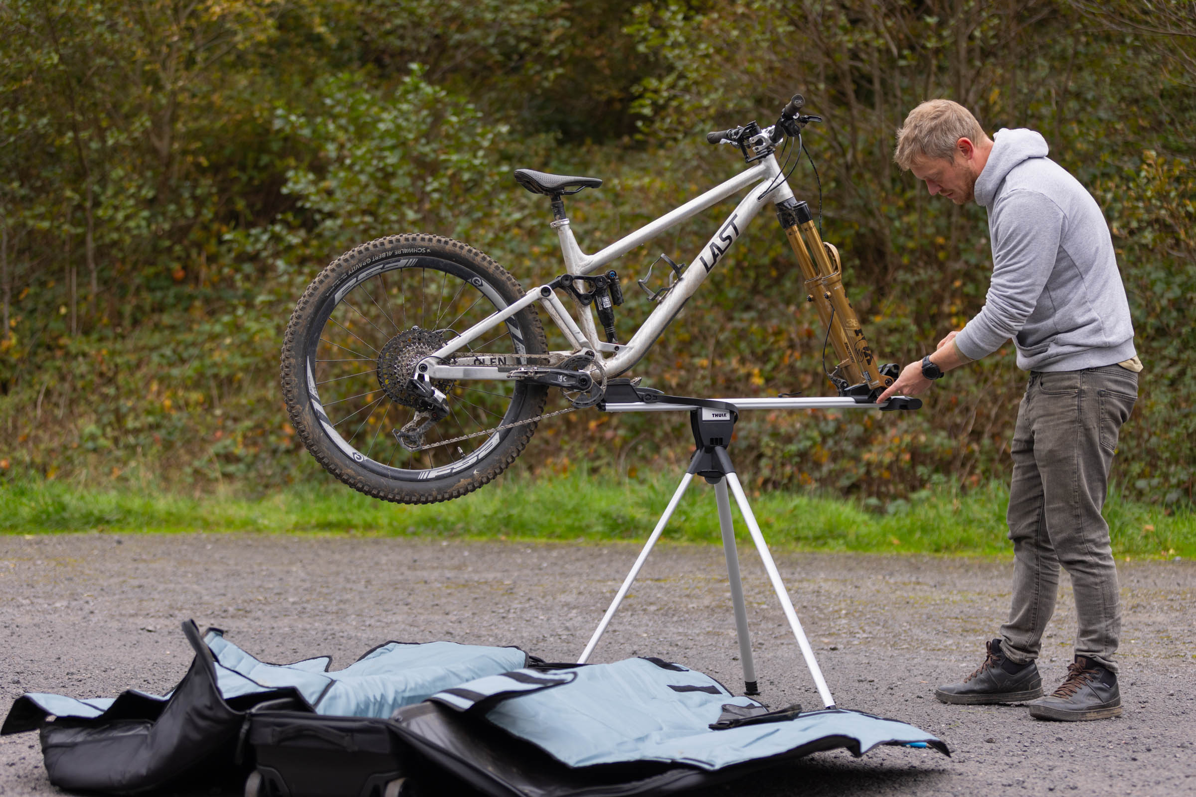 Person working on a silver Last full-suspension mountain bike mounted on the Thule RoundTrip MTB's three-legged workstand, with the travel case lying open in the foreground.