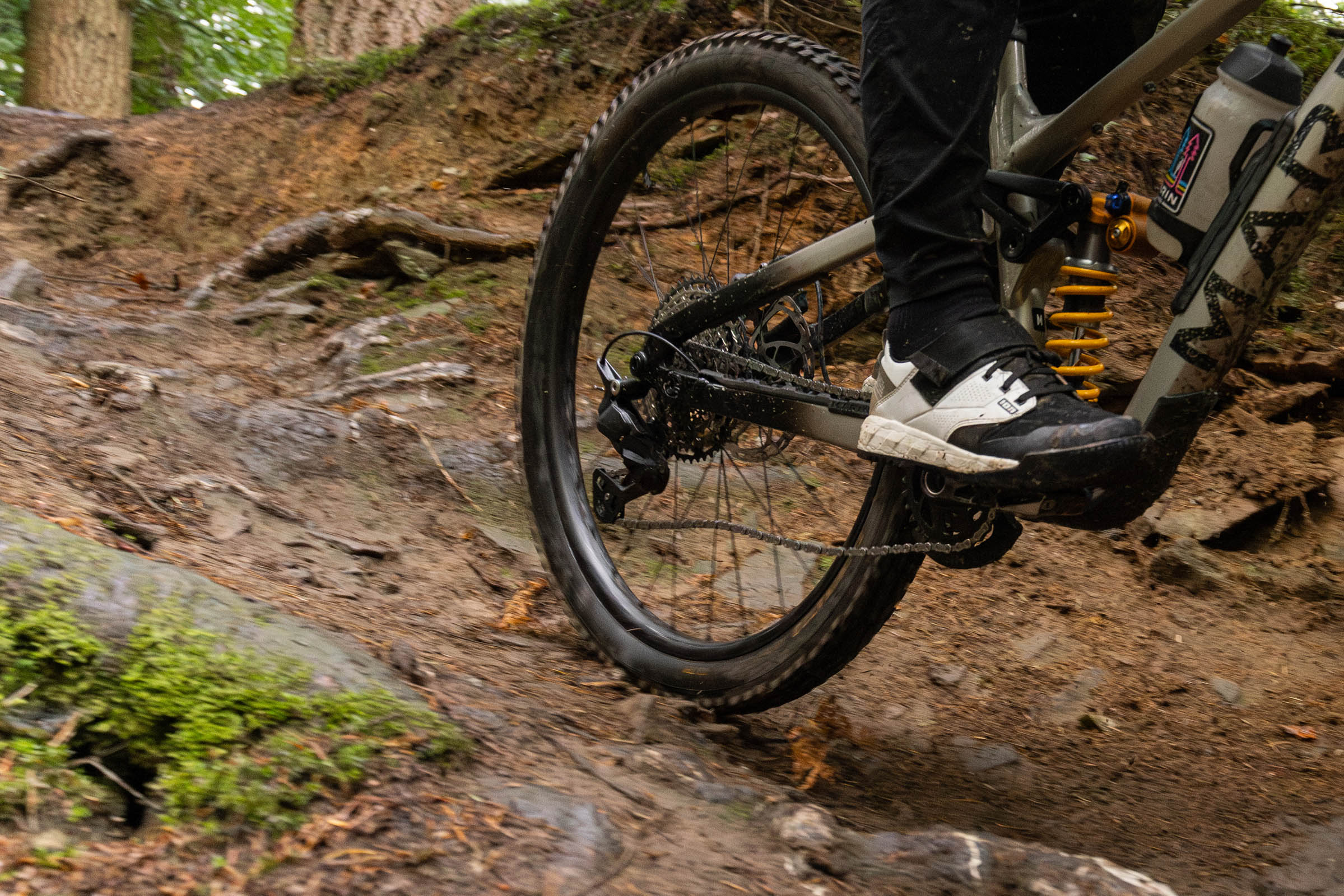 Close-up action shot of a mountain bike rear wheel and Shimano Deore M7200 drivetrain riding over rough roots and rocks.