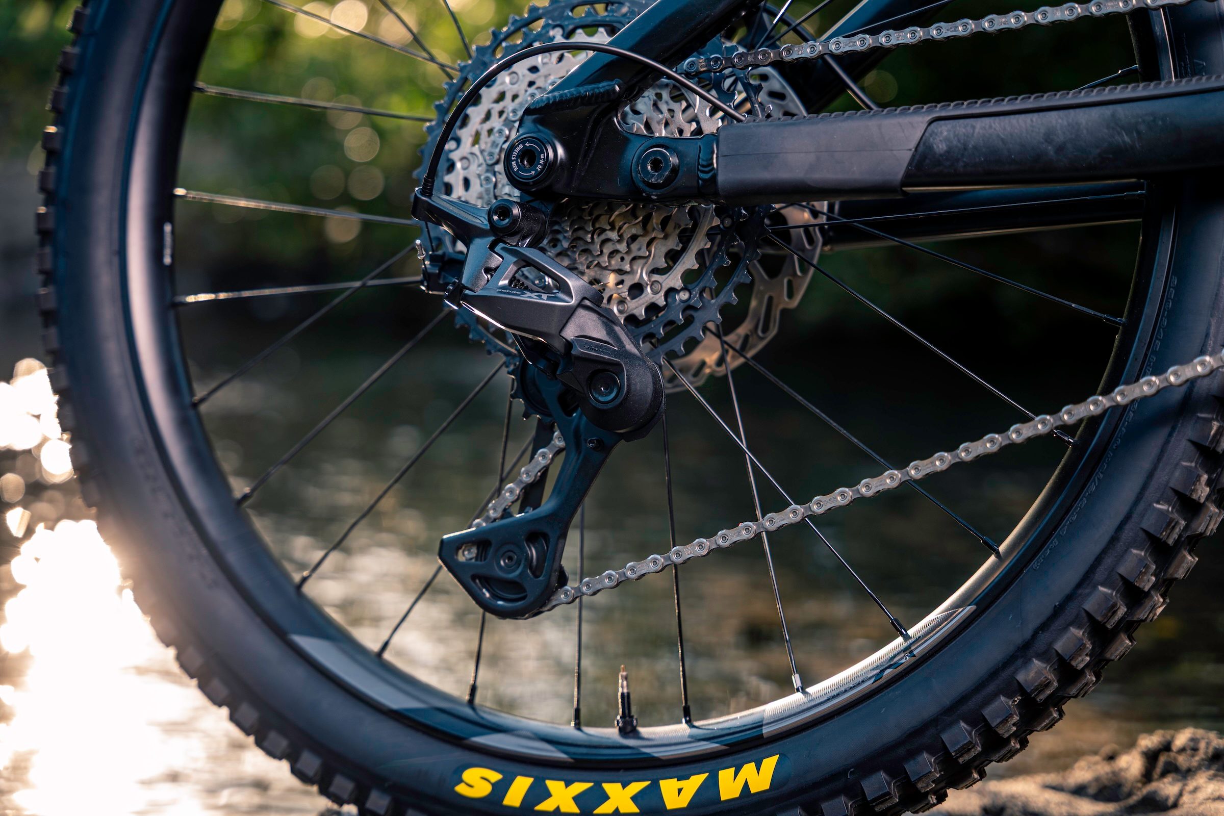 Close-up of a Shimano Deore XT M8200 rear derailleur and cassette fitted to a mountain bike beside water.