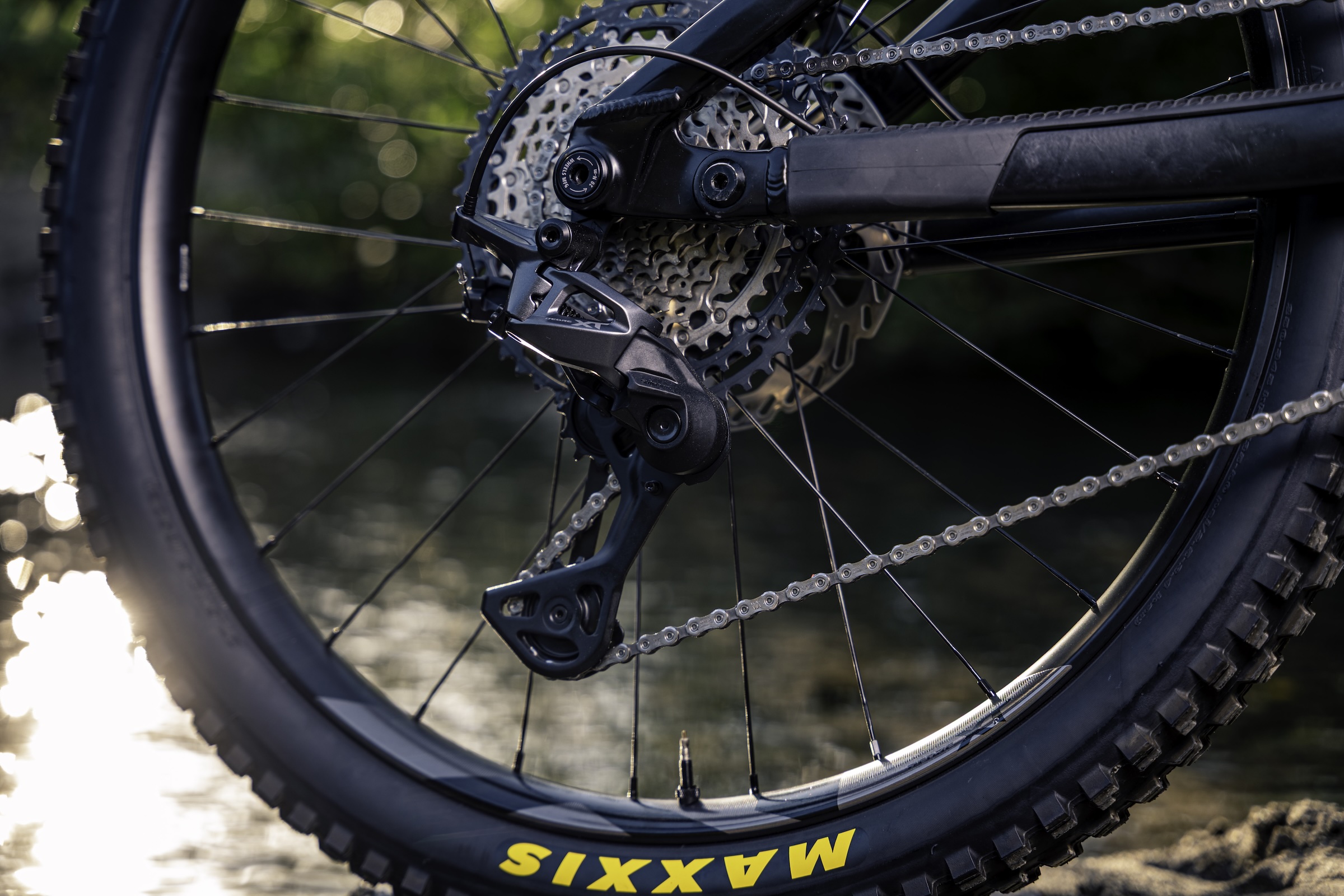 Close-up of a Shimano Deore XT M8200 rear derailleur and cassette fitted to a mountain bike beside water.